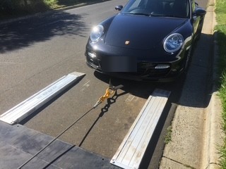 A black Porsche 911 being driven up the ramps onto the tilt tray, with a recovery strap attached